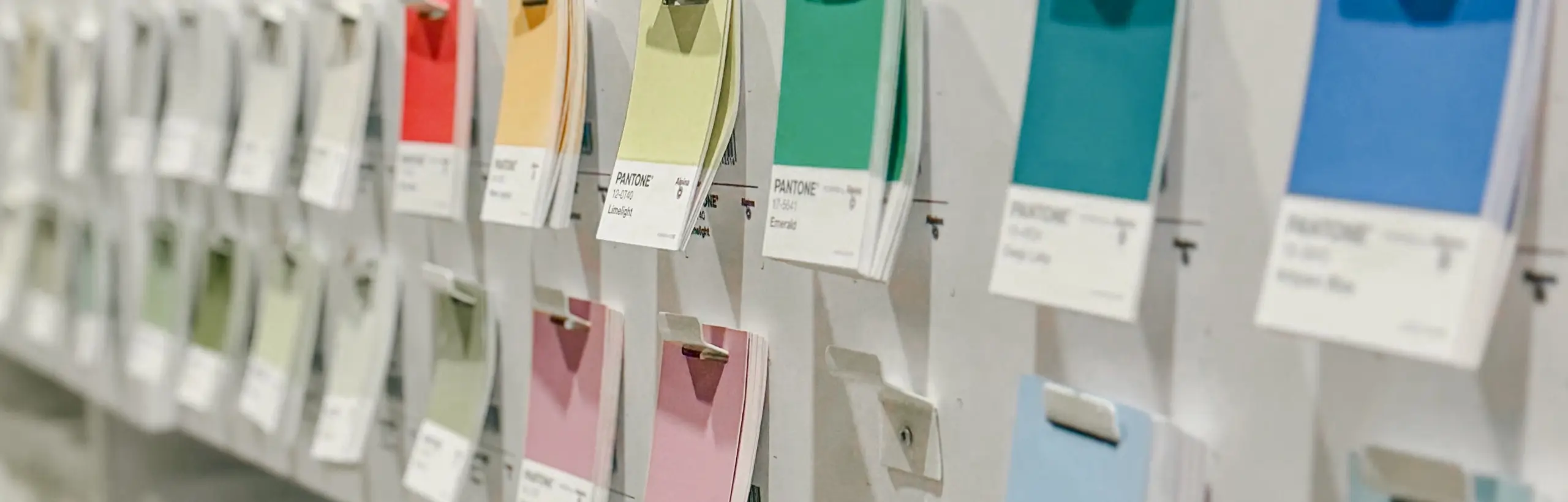 Aus schrägem Winkel ist ein Ausschnitt mehrfarbiger Pantone-Farbkärtchen zu sehen, die in zwei Reihen an der Leiste eines hellgrauen Stahlregals hängen.