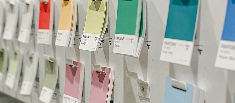 Aus schrägem Winkel ist ein Ausschnitt mehrfarbiger Pantone-Farbkärtchen zu sehen, die in zwei Reihen an der Leiste eines hellgrauen Stahlregals hängen.