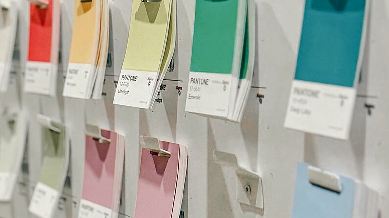 Aus schrägem Winkel ist ein Ausschnitt mehrfarbiger Pantone-Farbkärtchen zu sehen, die in zwei Reihen an der Leiste eines hellgrauen Stahlregals hängen.