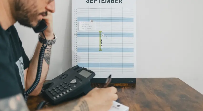 Eine Person sitzt an einem Tisch und hält den Hörer eines schwarzen, kabelgebundenen Telefons ans Ohr. An der Wand lehnen ein Monatskalender mit einer siebentägigen Markierung „CAMPING!“ sowie ein handgeschriebener Zettel mit der Aufschrift „TO-DO: Packliste, Route planen, Handtücher?“. Unter der rechten Hand, die einen Stift hält, liegt ein Notizblock. Die Nahaufnahme zeigt nur einen kleinen Ausschnitt der Person.