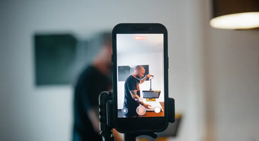 Ein Smartphone auf einem Stativ filmt eine Person beim Bedienen einer Textildruckpresse. Auf dem Display ist die Person scharf zu sehen, während dasselbe Motiv im Hintergrund unscharf erscheint.
