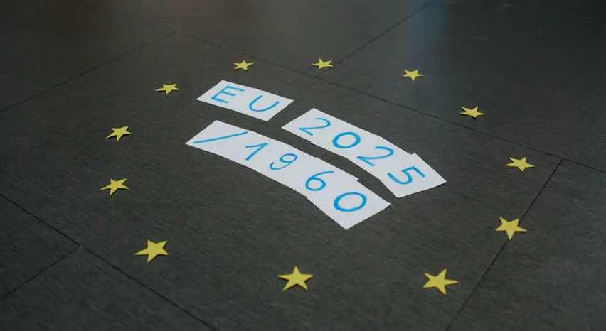 Auf einem grauen Steinboden liegen Zettel, auf denen in blauer Farbe die Zeichen „EU 2025/1960“ verteilt geschrieben stehen. Jeder Zettel trägt ein einzelnes Zeichen. Drumherum sind zwölf gelbe Papiersterne kreisförmig angeordnet, die das EU-Symbol darstellen. Das Foto ist aus einem schrägen, bodennahen Winkel aufgenommen.