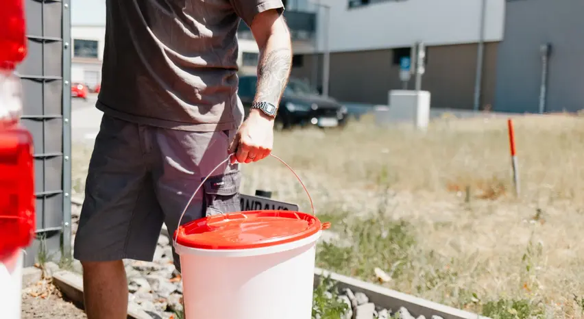 Ein Mann in brauner Handwerkerkleidung ist von der Brust bis zu den Waden zu sehen. Er steht mit dem Rücken zur Kamera vor dem Heck eines Kastenwagens und hält in der linken Hand einen weißen Farbeimer mit rotem Deckel. Im Hintergrund sind Steine, eine vertrocknete Wiese, ein Gebäude und ein geparktes Auto zu sehen.