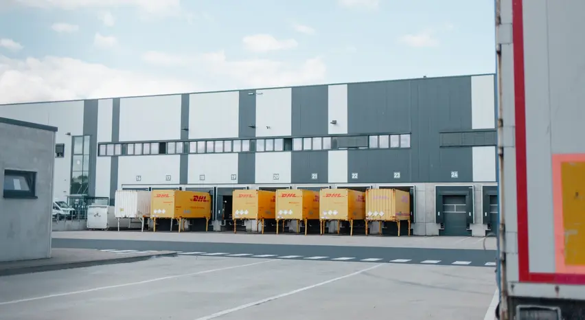 Mehrere Frachtcontainer stehen an einem länglichen, grauen Gebäude mit Rolltoren angedockt. Auf dem betonierten Boden sind Fahrbahnmarkierungen und Parkplätze eingezeichnet. Im Vordergrund sind am rechten Bildrand das Heck eines Lkw im Anschnitt und links ein kleinerer grauer Gebäudeblock ebenfalls im Anschnitt zu sehen.
