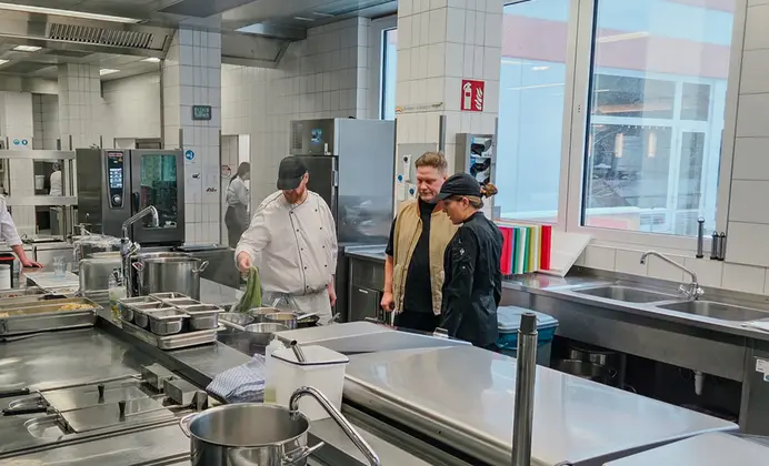 Ausschnitt einer Großküche mit glatten, silbernen Flächen. Die Wände sind weiß gefliest und mit Sicherheitszeichen beschildert. Neben einem Mann in weißer Kochuniform steht ein weiterer Mann in Alltagsbekleidung, neben ihm eine Frau in schwarzer Küchenhilfenuniform. Die Blicke sind auf die Hand des Kochs gerichtet.