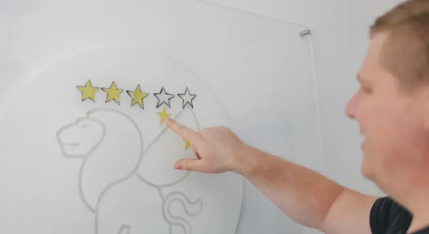 Im Anschnitt ist ein transparentes Glas-Whiteboard mit einem mittig platzierten weißen Gandayo-Logo zu sehen. Auf dem Board sind die Umrisse von fünf Sternen dargestellt. Ein Mann schiebt mit dem Zeigefinger gelbe Papiersterne in die Umrisse. Sein Gesicht ist nur unscharf am rechten Bildrand zu erkennen.