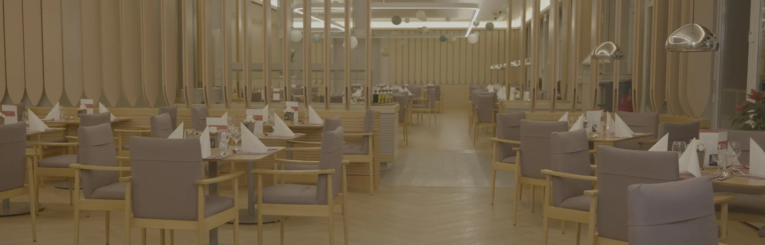 Ein leerer Speisesaal eines Restaurants. Der Raum ist in zartes, helles Licht getaucht und stilvoll mit hölzernen Elementen und silbernen Leuchten eingerichtet. Graue Polsterstühle stehen um gedeckte Tische mit aufgestellten Stoffservietten.