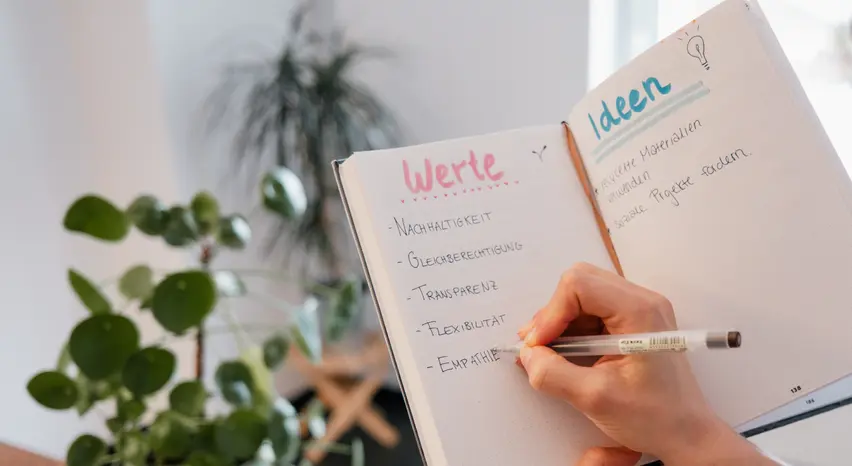 Nahaufnahme eines aufgeschlagenen Notizbuchs, welches in der Hand gehalten wird. Die zweite Hand schreibt auf die linke Seite des Punktraster-Papiers Werte wie „Nachhaltigkeit, Gleichberechtigung, Transparenz, Flexibilität und Empathie“ und auf die rechte Seite Ideen wie „recycelte Materialien verwenden“ und „soziale Projekte fördern“.