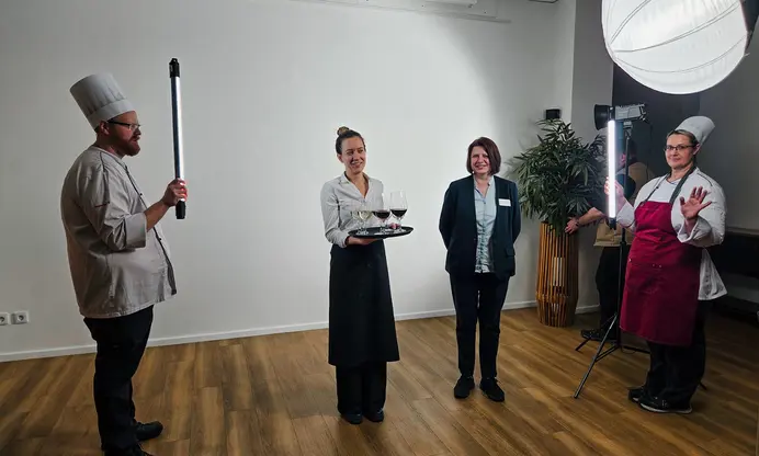 Vier Personen stehen in einem Raum mit Holzparkett und weißer Wand. Links außen steht ein Mann, rechts außen eine Frau, die jeweils eine Kochuniform tragen und eine Leuchtröhre auf die beiden Frauen in der Mitte halten. Die Köchin rechts außen winkt in die Kamera, hinter ihr ist ein Stativ mit Kamera zu sehen, über ihr eine Kugelleuchte. Die linke der beiden mittleren Personen trägt eine Kelleruniform und vor sich ein Tablett mit mehreren gefüllten Weingläsern. Die Frau daneben trägt einen schwarzen Hosenanzug, ein hellblaues Hemd und ein Namensschild. Sie lächelt in die Kamera.