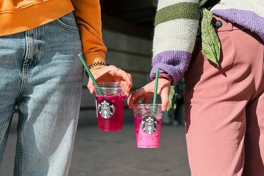 Nahaufnahme von zwei nebeneinanderstehenden Personen, die Plastikbecher mit Starbucks-Aufdruck auf Höhe ihrer Beine in ihren Händen halten. In den Bechern sind rosafarbene Getränke.  