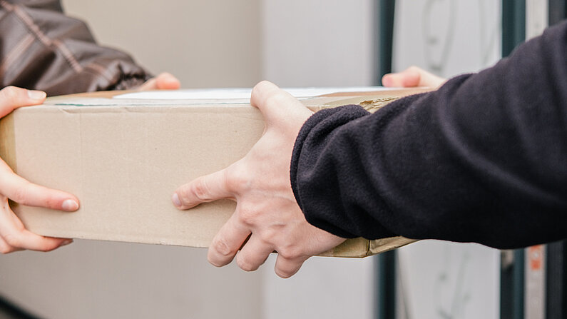 Nahaufnahme einer Paketübergabe. Zwei einander gegenüberstehende Personen überreichen sich einen Karton. 