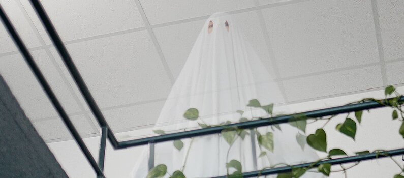 Eine als Geist verkleidete Person steht oben hinter dem Geländer eines Treppenhauses, welches von rankenden Pflanzen bewachsen ist.