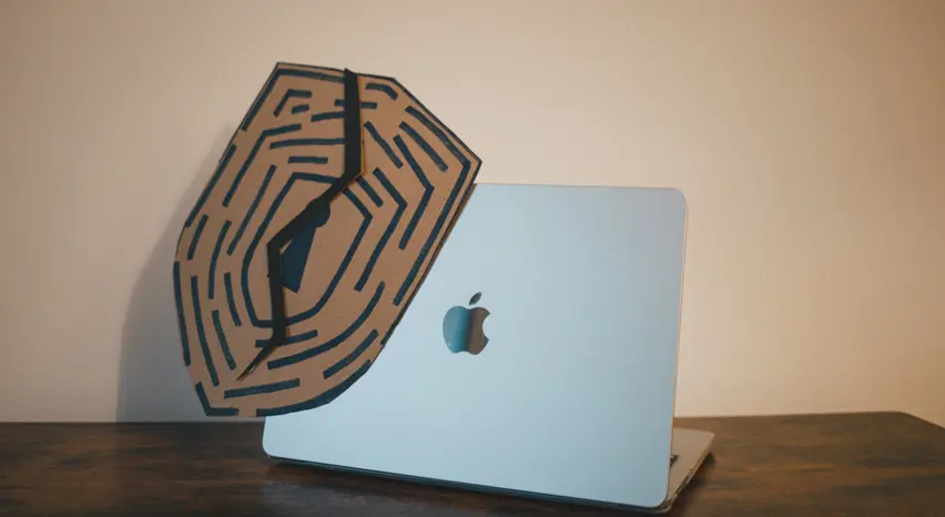 Die Rückansicht eines aufgeklappten MacBooks zeigt ein diagonal an der oberen linken Ecke angebrachtes Schutzschild aus Pappe, das von einer schwarzen, gezackten Linie durchtrennt wird.