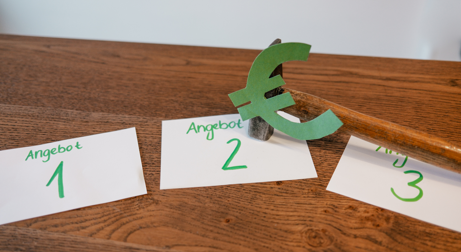 Auf einem Tisch liegen drei Zettel mit der grünen Aufschrift „Angebot 1/2/3“. Ein Hammer, auf dessen Kopf ein großes grünes Euro-Zeichen aus Papier geklebt ist, liegt auf dem mittleren Zettel „Angebot 2“. 