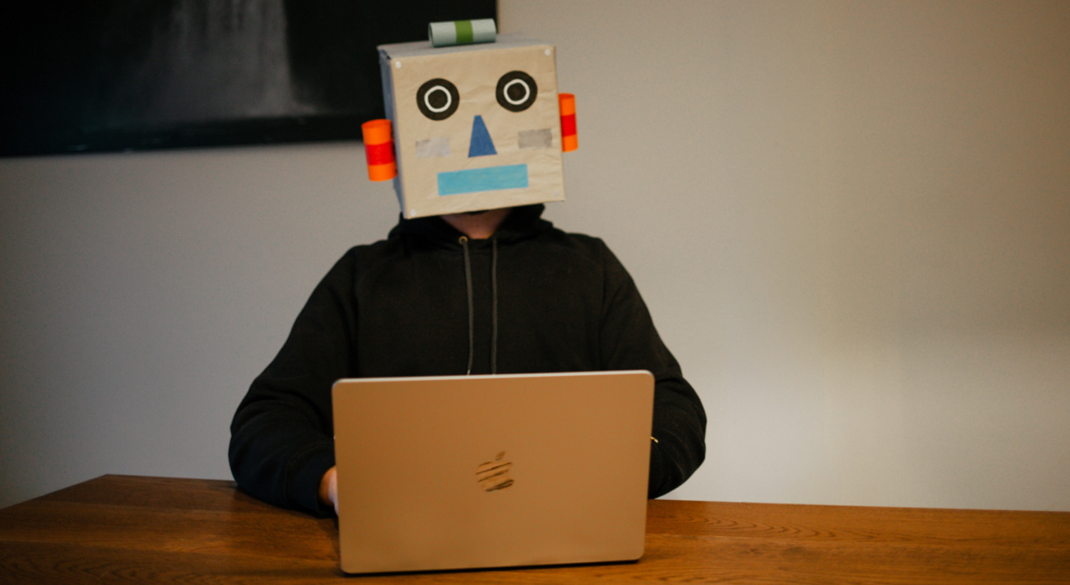 Vor einem aufgeklappten MacBook sitzt eine Person in einem schwarzen Hoodie. Ihr Kopf ist von einem selbst gebastelten Roboterkopf aus Pappe verdeckt.