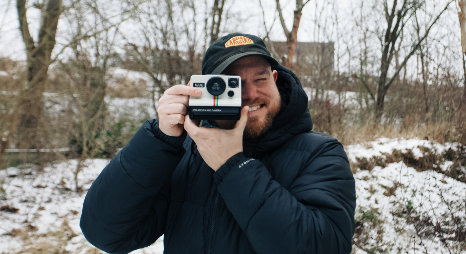 Ein lächelnder Mann mit schwarzer Kappe und dunkelblauer Winterjacke schaut durch eine Polaroid Land Camera 1000 und hat den Finger auf dem Auslöser platziert. Im Hintergrund stehen kahle Bäume auf leicht mit Schnee bedeckter Erde.
